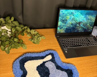 May include: A blue, white, and black rug with a wavy, abstract design. The rug is on a wooden surface next to a laptop.