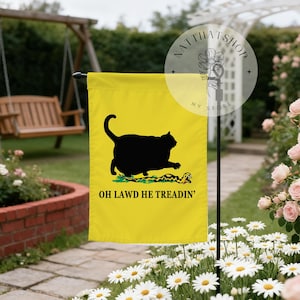 May include: A yellow garden flag features a black cat silhouette stepping on a snake, with the text "OH LAWD HE TREADIN'". The flag is displayed outdoors, surrounded by flowers and greenery.