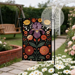 May include: A black garden flag with a floral design, featuring a large purple iris at the centre. The design includes orange, yellow, and red flowers with green leaves. The flag is displayed outdoors in a garden setting.