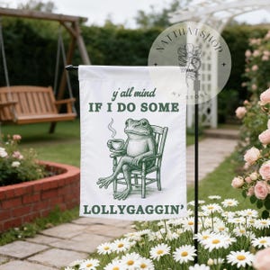 May include: White garden flag featuring a green frog sitting in a chair, holding a cup of coffee. The flag has the text "y'all mind IF I DO SOME LOLLYGAGGIN'" in green. The flag is displayed outdoors in a garden setting.