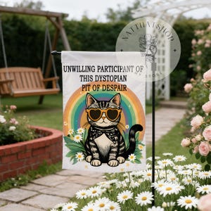 May include: A white garden flag with a cat wearing heart-shaped sunglasses, a bow tie, and a rainbow. The text reads "UNWILLING PARTICIPANT OF THIS DYSTOPIAN PIT OF DESPAIR". The background shows daisies and a garden setting.