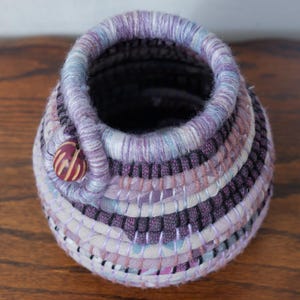 May include: A small, hand-woven basket in shades of purple, lavender, and white. The basket has a button closure and a rounded shape. The woven design creates a textured surface, ideal for home decor.