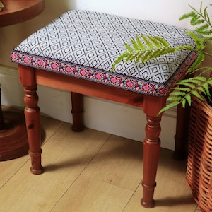 May include: A small wooden stool with a patterned cushion. The cushion features a black and white geometric design with a colourful border. The stool has four turned legs and is made of stained wood. A fern and a wicker basket are in the background.