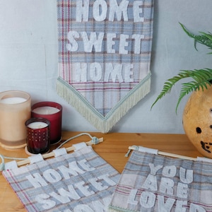 May include: Decorative fabric banners with the phrases "HOME SWEET HOME" and "YOU ARE LOVED" in white lettering on a plaid design. Each banner has a wooden dowel and fringe. Candles and a vase are also visible.
