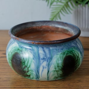 May include: A round ceramic planter with a blue, green, and white marbled design. The rim is dark blue and brown. The interior is a reddish-brown colour. The planter sits on a wooden surface.