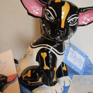 May include: A ceramic dog figurine with a glossy black finish, adorned with white and yellow bone accents. The dog has large, pink-lined ears and a tag that reads "WhimsiClay". The figurine is sitting on a blue surface.