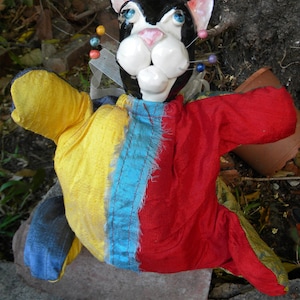 May include: A black and white cat puppet with a red hat and a colorful patchwork costume. The puppet has a long, thin nose and large, round eyes. The costume is made of a variety of fabrics, including yellow, red, blue, and green.