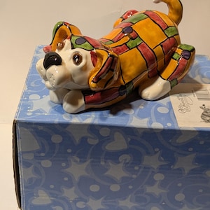 May include: A ceramic dog figurine in a playful pose, featuring a colorful stained-glass design. The dog has a white face, black nose, and brown eyes. The body is a patchwork of yellow, red, green, and blue. The figurine is on a blue box.