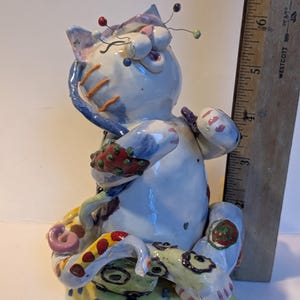 Puede incluir: Una figura de gato de cerámica con cuerpo azul claro, detalles estampados coloridos y varios alfileres de costura insertados en la cabeza. El gato se sienta sobre una base amarilla con lunares junto a una regla de madera de 18 cm.