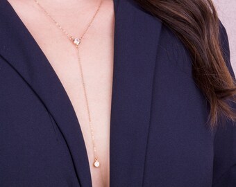 May include: A delicate gold-tone necklace with a Y-shaped design. The necklace features a small, round, clear gemstone at the top and a larger, similar gemstone at the bottom. The necklace is worn over a navy blue blazer.
