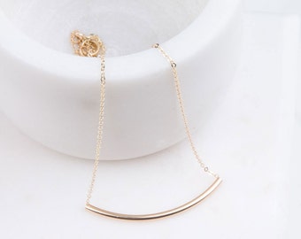 Puede incluir: Un collar de oro con un colgante de barra curvada. El collar es delicado y minimalista.