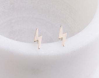 Puede incluir: Un par de pequeños pendientes de plata en forma de rayo. Los pendientes están sobre una superficie blanca.