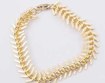 Könnte beinhalten: Ein goldenes Kettenarmband mit einem Chevron-Muster. Die Kette besteht aus kleinen, spitzen Gliedern, die ein durchgehendes, sich wiederholendes Muster bilden.
