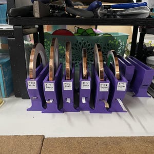 Pode incluir: Bobinas de fita de cobre em suportes roxos. As bobinas de fita são rotuladas com tamanhos e materiais como prata, preto e cobre. A imagem mostra uma configuração de suprimentos de artesanato, com ferramentas e outros materiais.