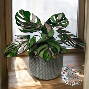 May include: Stained glass Monstera plant with green, white, and pink leaves in a grey textured pot. The plant is displayed on a wooden windowsill. The image is of a decorative item.