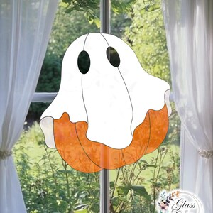 Op de afbeelding: Een gebrandschilderde Halloween-decoratie met een spookontwerp. Het spook heeft een witte body met zwarte ovale ogen en een oranje pompoenvormige basis. De decoratie hangt voor een raam met witte gordijnen en een groen uitzicht.