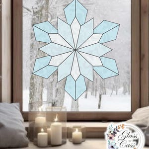May include: A stained glass snowflake in shades of blue and white is displayed in a window. The snowflake is composed of geometric shapes and is set against a snowy winter landscape. Candles in glass holders sit on the windowsill.