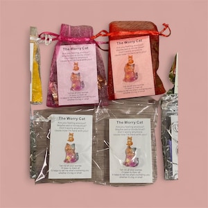 May include: Several small, decorative cat figurines in pink and red organza bags, and clear plastic bags. Each bag has a card that reads "The Worry Cat" with a poem and a cat illustration. The background is a light pink.