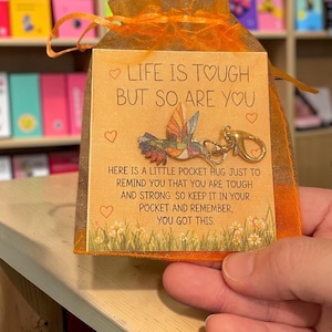 May include: An orange organza drawstring bag containing a square card with a colorful hummingbird charm and gold lobster clasp. The card features the text: LIFE IS TOUGH BUT SO, ARE YOU. HERE IS A LITTLE POCKET HUG JUST TO REMIND YOU THAT YOU ARE TOUGH AND STRONG. SO KEEP IT IN YOUR POCKET AND REMEMBER, YOU GOT THIS.