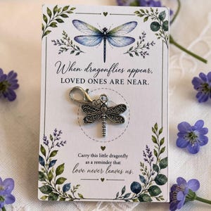 May include: A silver-toned metal dragonfly charm with a lobster clasp attached to a white rectangular card. The card features a watercolor dragonfly illustration, green foliage, and the text: When dragonflies appear, LOVED ONES ARE NEAR.