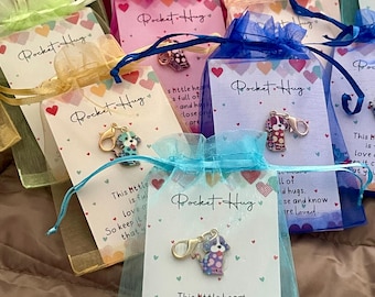 May include: A collection of small gift bags in various colors, each containing a charm shaped like a dog. The bags have a tag that reads "Pocket Hug" and a message about love and hugs. The charms are attached to a clasp.
