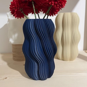 Könnte beinhalten: Drei dekorative Vasen mit unterschiedlichen Designs. Die vordere Vase ist marineblau und enthält rote Blumen. Die anderen Vasen sind weiß und beige. Die Vasen haben ein wellenförmiges, strukturiertes Design.