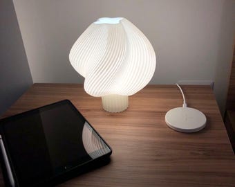 Könnte beinhalten: Eine weiße, moderne Tischlampe mit einem einzigartigen, wirbelnden Schirmdesign. Die Lampe steht auf einem Nachttisch aus Holz, neben einem schwarzen Tablet und einem weißen kabellosen Ladegerät. Der Lampenschirm hat eine gerippte Textur.