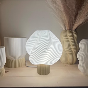 Può includere: Una lampada da tavolo bianca, strutturata, con un esclusivo design a spirale del paralume. La lampada poggia su una base beige ed è illuminata. Sono visibili anche altri vasi decorativi, che creano un tema di arredamento moderno.