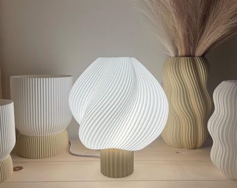 Könnte beinhalten: Eine weiße, strukturierte Tischlampe mit einem einzigartigen, wirbelnden Schirmdesign. Die Lampe steht auf einem beigen Sockel und ist beleuchtet. Weitere dekorative Vasen sind ebenfalls zu sehen, die ein modernes Wohn-Thema schaffen.
