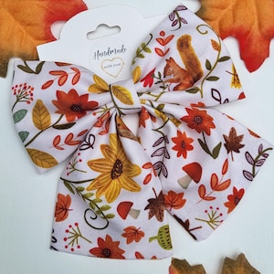 May include: A white fabric hair bow with a fall-themed pattern featuring orange and yellow flowers, leaves, mushrooms, and a squirrel. The bow is labeled "Handmade with love".