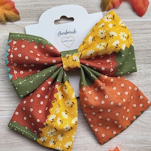 May include: A handmade fabric bow with a patchwork design. The bow features sections of orange, yellow, and green fabric, each with a different floral or dotted pattern. The bow is attached to a white tag with the words "Handmade with love".