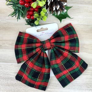 May include: A large hair bow featuring a red, green, and black plaid pattern with a gold thread accent. The bow is attached to a white display card printed with the text Twinkle Bloom Boutique and a small butterfly icon.