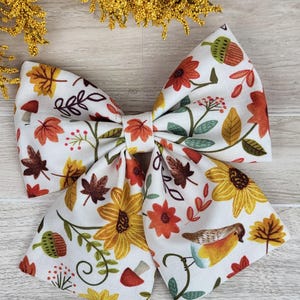 May include: A white fabric hair bow featuring a repeating autumn-themed pattern of yellow sunflowers, orange and brown maple leaves, small red mushrooms, green acorns, and a small bird with a yellow chest and blue wing accents.
