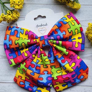 May include: A large, handmade bow with a colorful jigsaw puzzle pattern. The bow features a variety of bright colors, including red, blue, green, yellow, and purple. The bow is attached to a white tag that reads "Handmade with love."