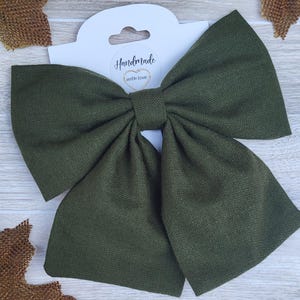 May include: An olive green fabric bow with a large, symmetrical design. The bow is attached to a white tag with the words "Handmade with love" in cursive script. The bow is made of a textured material.