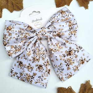 May include: A large, handmade hair bow with a floral pattern in shades of brown and tan on a white background. The bow is attached to a white tag with the words "Handmade with love."