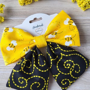 May include: A hair bow featuring two distinct fabric patterns. The top section is bright yellow with a repeating pattern of small bees and white wings. The bottom section is black with a yellow dashed line swirl pattern. A white tag reads Handmade with love.