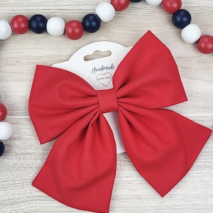 May include: A large, bright red fabric bow on a white tag with the text "Handmade with love". The bow is centered on a light wood grain surface, with a beaded garland of red, white, and blue beads in the background.