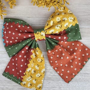 May include: A fabric hair bow featuring a patchwork design of various floral patterns in shades of mustard yellow, forest green, and terracotta red. The bow is constructed from multiple segments of cotton-like fabric with small white flower prints.