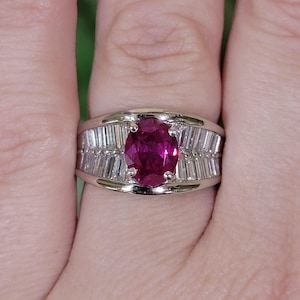 May include: A silver-toned ring featuring a central oval-cut deep pink gemstone held by four prongs. The band is flanked on both sides by a row of clear, rectangular baguette-cut stones set horizontally within the polished metal band structure.