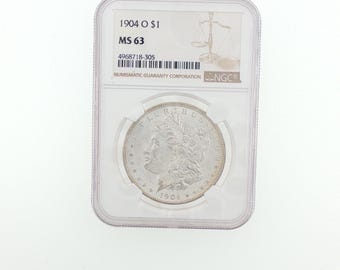 Op de afbeelding: Een zilveren Morgan dollar munt uit 1904 O, ingesloten in een doorzichtige plastic NGC-houder. Het label toont de tekst 1904 O $1, MS 63 en het serienummer 4968718-305. De munt toont het profiel van Lady Liberty met sterren.
