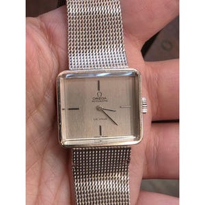 Pode incluir: Um relógio de pulso automático Omega De Ville vintage com mostrador retangular dourado e pulseira de malha metálica. O mostrador exibe o logotipo da Omega, as palavras "AUTOMATIC" e "DE VILLE" e as palavras "SWISS MADE".