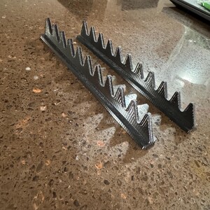 May include: Two black plastic 3D printed shelf dividers with a zig-zag design. The dividers are on a brown countertop.
