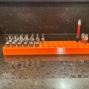 May include: An orange rectangular bit holder with multiple holes, holding various silver and red screwdriver bits. The holder is made of plastic and is designed to organize and store different types of screwdriver bits.