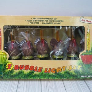 May include: A 7 bubble light set in a tan cardboard box featuring turkey-shaped bases. The box displays the text 7 BUBBLE LIGHT SET, See Them Bubble!, and UL LISTED. The packaging shows illustrations of green and red candle-style bubble lights.