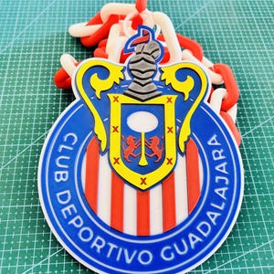 May include: A round, blue, red, and white Club Deportivo Guadalajara emblem. The emblem features the team's name and a shield with a knight's helmet and two red lions. A chain of red and white links is attached to the emblem.