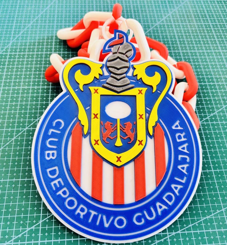 May include: A round, blue, red, and white Club Deportivo Guadalajara emblem. The emblem features the team's name and a shield with a knight's helmet and two red lions. A chain of red and white links is attached to the emblem.