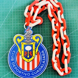 May include: A decorative pendant featuring the Club Deportivo Guadalajara logo, with a red, white, and blue color scheme. The pendant is connected to a chain of alternating red and white links. The logo is a shield shape with a crest.