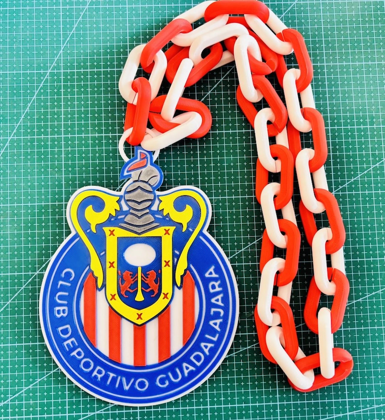 May include: A decorative pendant featuring the Club Deportivo Guadalajara logo, with a red, white, and blue color scheme. The pendant is connected to a chain of alternating red and white links. The logo is a shield shape with a crest.