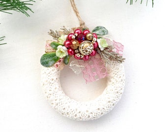 May include: White crocheted wreath ornament with a pink lace bow and red berry accents. The wreath is decorated with gold glitter and beads.
